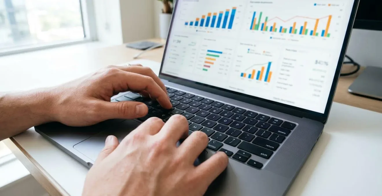 Plano detalle de manos tecleando en teclado de ordenador portátil moderno con pantalla borrosa mostrando gráficos de datos en segundo plano sobre superficie limpia