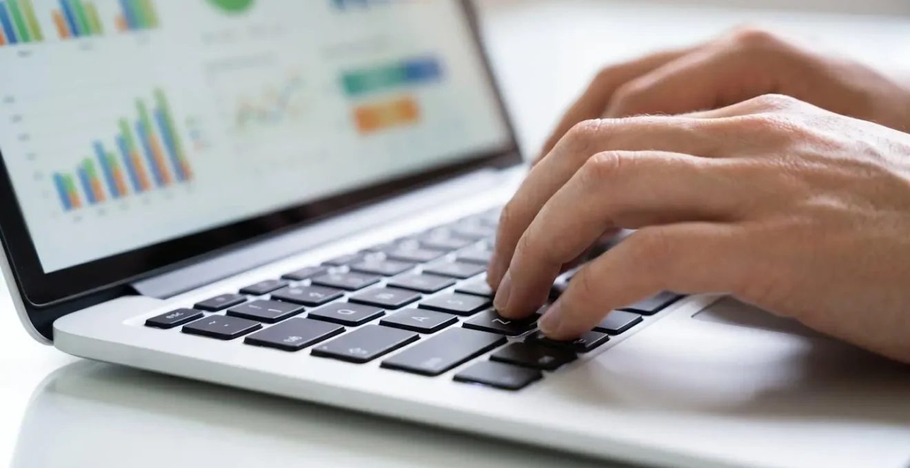 Plano detalle de manos tecleando en teclado de ordenador portátil moderno con pantalla borrosa mostrando gráficos de datos en segundo plano sobre superficie limpia